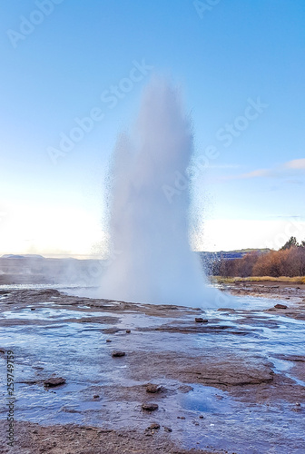 The Great Geysir, active geyser. Hot spring in a geothermal area. Steam of water bursts high up in the sky, splashing water all around. Water flowing on the rocks. Clear blue sky.