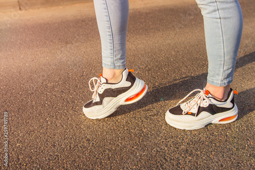 Girl in sneakers and jeans. Several pairs of sports shoes and legs. Stylish fashionable white women s leather sneakers on asphalt on a sunny day. Female legs with sneakers