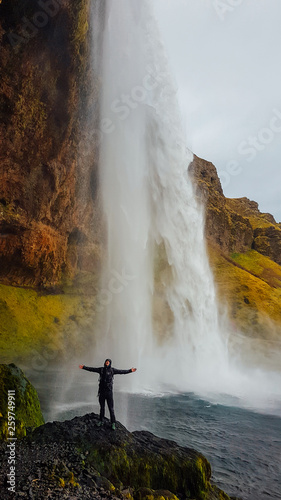 A young man stands in front of a waterfall, with his arms spread open. He is overjoyed and happy. Huge and powerful waterfall. Slopes of the mountain overgrown with moss and golden grass. Being close
