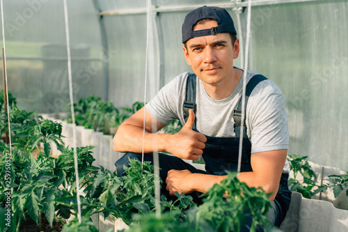 Handsome cheerful young gardener in overall and cap working in greenhouse. Portrait of joyful farmer.