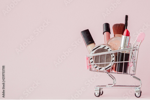 Creative concept with shopping trolley with makeup on a pink background. Perfume, sponge, brush, mascara, pencil, nail file, eye shadow, lip gloss in the basket, copy space photo