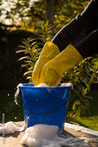 Spring Cleaning outside with big yellow cleaning gloves, water, soap and a big blue bucket with soap. Cleaning the table. Soap Foam on the table. Hands in bucket. Holding and wringing cleaning sponge.