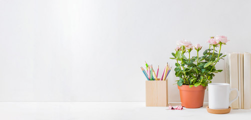 Home office, interior for bloggers workplace with roses in a pot, books, white mug, office supplies