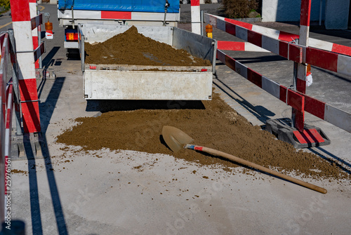 Pipeline construction site, with open tailgate and sand with shovel on the street. Trailer is loaded with sand. Striped barricades close temporary the road construction and keeps danger outside.