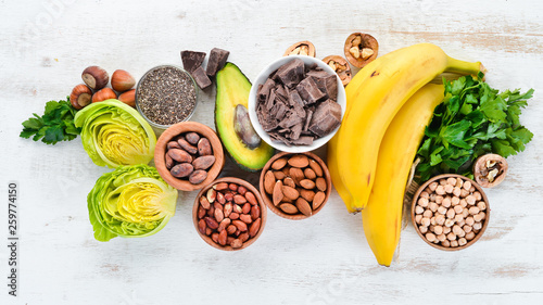 Foods containing natural magnesium. Mg: Chocolate, banana, cocoa, nuts, avocados, broccoli, almonds. Top view. On a white wooden background. photo