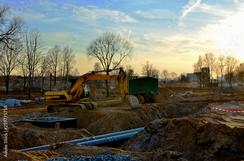 Tracked excavator with a bucket in the ground at a construction site during the laying of sewer pipes in the ground  against a blue sky and sunset