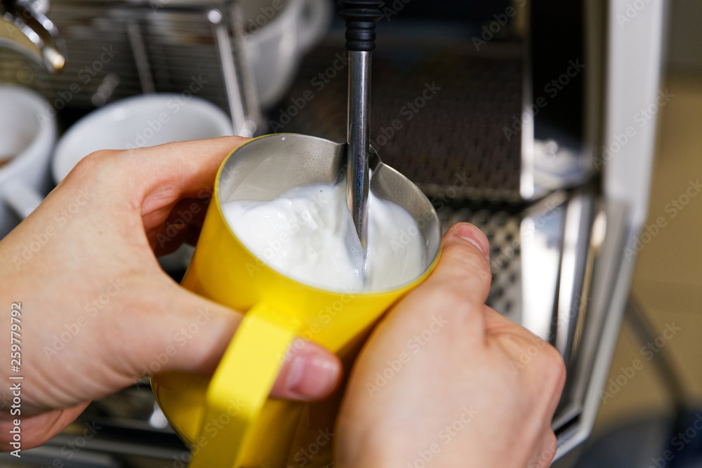 Closeup hands of barista who steaming milk for cappuccino or latte in yellow pitcher.