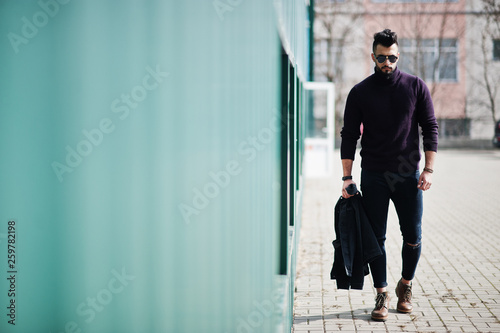 Fashion rich beard Arab man wear on turtle neck and sunglasses walking against green wall with jacket on hand. Stylish, succesful and fashionable arabian model guy.