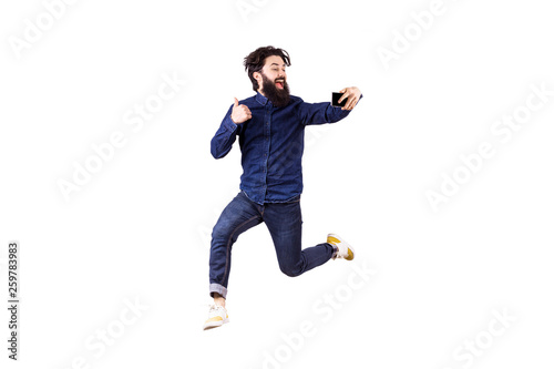 Full length portrait of a joyful bearded hipster man in glasses, jumping and taking selfie, isolated on white background