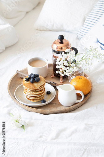Breakfast in bed. Pancakes with blueberries and honey, coffee, milk and fresh orange served on wooden tray on white sheets.