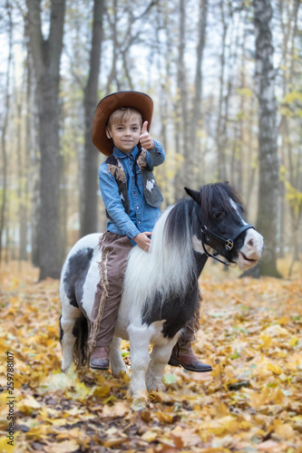 The boy in a suit of the cowboy sits astride a tiny pony in the park in the fall. The guy with a happy look, raised a finger up, expressing pleasure