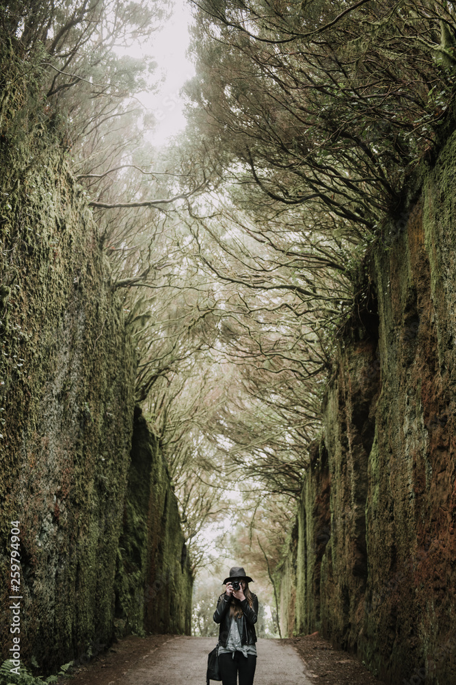 Elegant lady in hat and leather jacket shooting on camera and standing on footpath between murk alley of high walls and woods Elegant lady in hat and leather jacket shooting on camera and standing on footpath between murk alley of high walls and woods