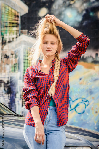 Portrait of a cheerful attractive blonde girl with a long braid, adjusting her hair, in a red shirt and jeans.