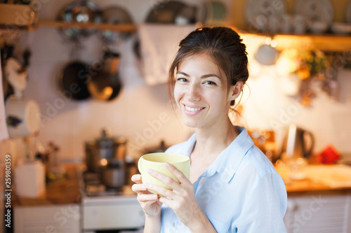 Young woman is drinking tea in cozy home kitchen. Happy girl is enjoying break in cooking and holding mug of coffee.