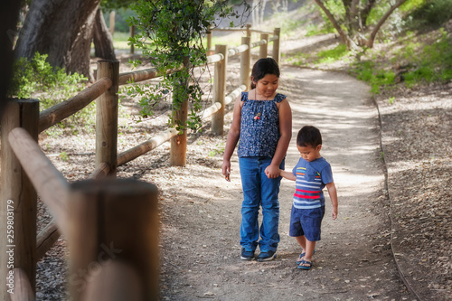 Older sister holding little brothers hand while walking in a wooded pathway in a California national park.