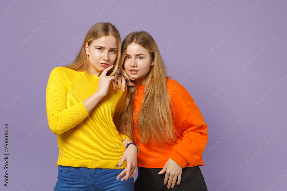 Two charming young blonde twins sisters girls in vivid colorful clothes standing, looking camera isolated on pastel violet blue wall background. People family lifestyle concept. Mock up copy space.
