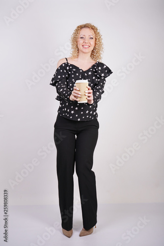 cute curly blonde girl stands in black fashionable clothes and holds a paper Cup of coffee on a white Studio background