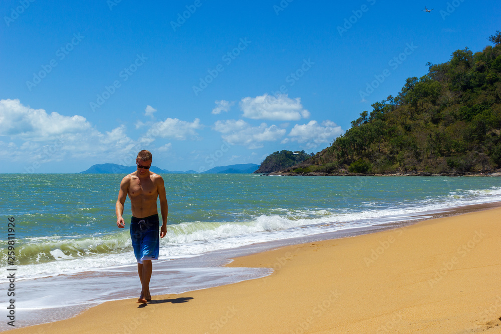 Muscular caucasian man walking on the beach and smiles as ocean waves crash behind him, cape tribulation, queensland, australia