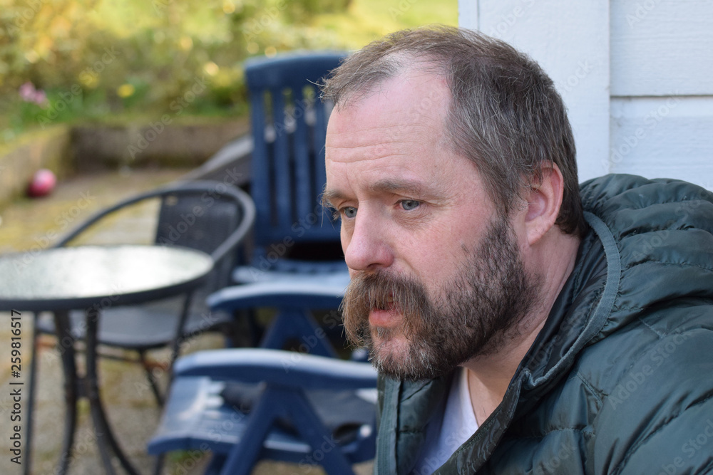 A bearded man sits against the background of garden furniture in the yard and looks thoughtfully. Blurred background. Close-up A bearded man sits against the background of garden furniture in the yard and looks thoughtfully. Blurred background. Close-up