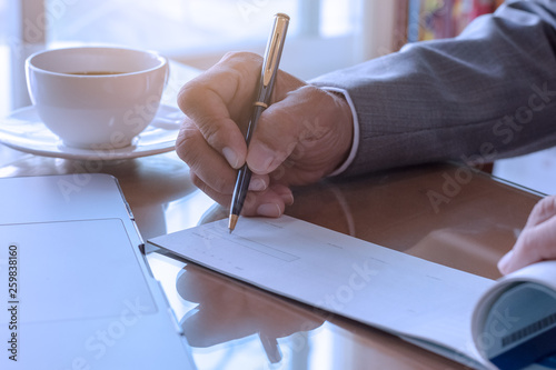 Business man hand writing and signing checkbook with laptop computer and white cup of coffee on the wooden table at modern home office.	 photo