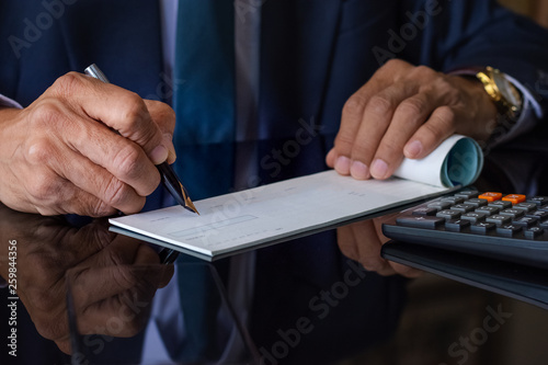 Businessman  in black suit with necktie, hand writing and signing checkbook on the wooden table at office. Paycheck or payment by cheque concept. photo