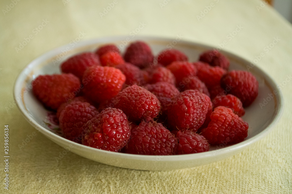Ripe raspberries are laying on the white plate which is standing on the table covered with yellow tablecloth Ripe raspberries are laying on the white plate which is standing on the table covered with yellow tablecloth