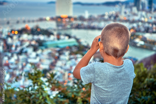 Young boy looking with the binoculars on the sunset city from the mountain viewpoint
