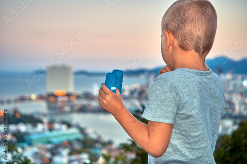 Young boy looking with the binoculars on the sunset city from the mountain viewpoint