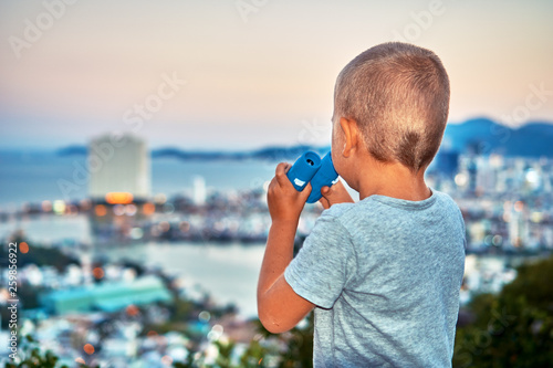 Young boy looking with the binoculars on the sunset city from the mountain viewpoint