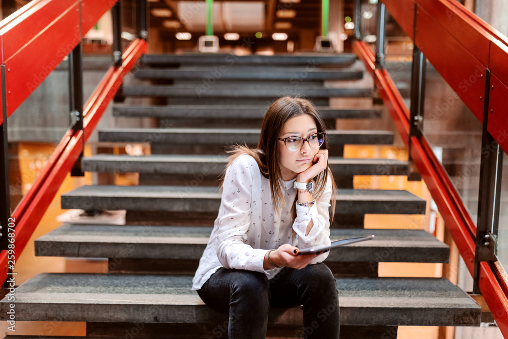 Fototapeta premium Bored collage girl with brown hair and eyeglasses leaning on the hand and sitting on the stairs and holding tablet. University building interior.