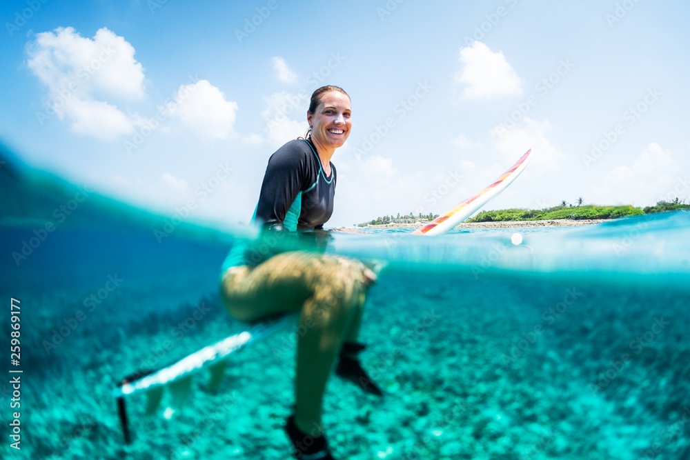Naklejka premium Happy lady surfer sits on the surfboard, smiles and looks at the camera . Splitted image with underwater view