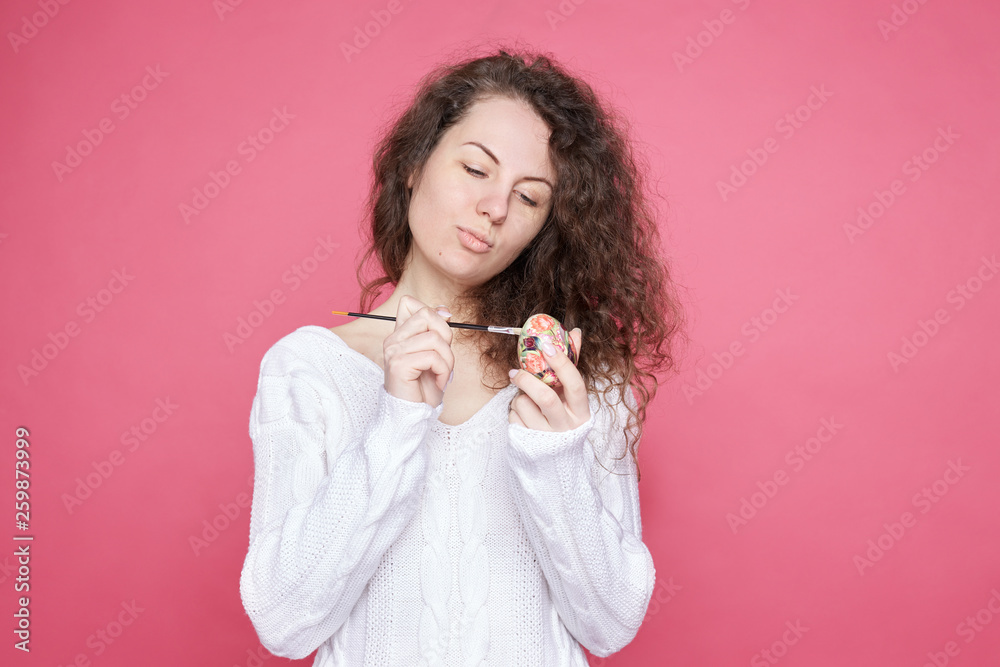 Concentrated curly haired lady holds traditional egg in hand, brush in other, painting it in different colors, rejoices coming holiday, wears cozy knitted outfit. Easter and spring celebration concept Concentrated curly haired lady holds traditional egg in hand, brush in other, painting it in different colors, rejoices coming holiday, wears cozy knitted outfit. Easter and spring celebration concept
