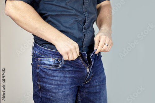 Man unzipps blue jeans. Man torso, fingerring, blue shirt, casual wear. Indoors, grey empty background, copy space.