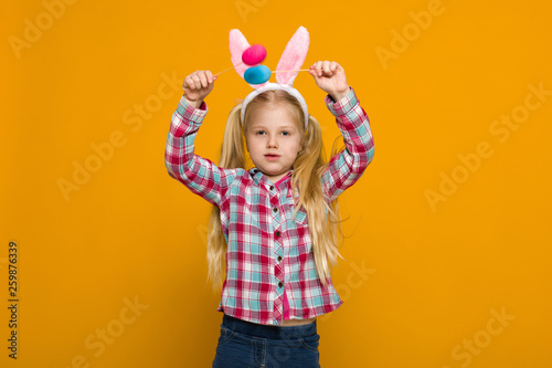 Portrait of cute little child girl with Easter bunny ears holding colorful eggs on yellow background. Happy easter