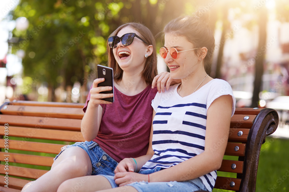 Loyal happy friends feel good near each other in the park. Beautiful cheerful girls read funny unpredictable story on bench burst out laughing loudly and have rest together on summer weekend.
