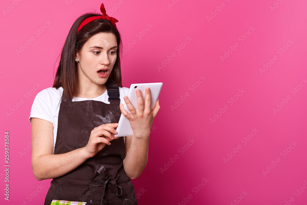 Shocked yound woman stands and watches breaking news on the white tablet,looks extremely astonished watching with widely open mouth. Sincere female wears work clothing, looks messy with flour. Shocked yound woman stands and watches breaking news on the white tablet,looks extremely astonished watching with widely open mouth. Sincere female wears work clothing, looks messy with flour.