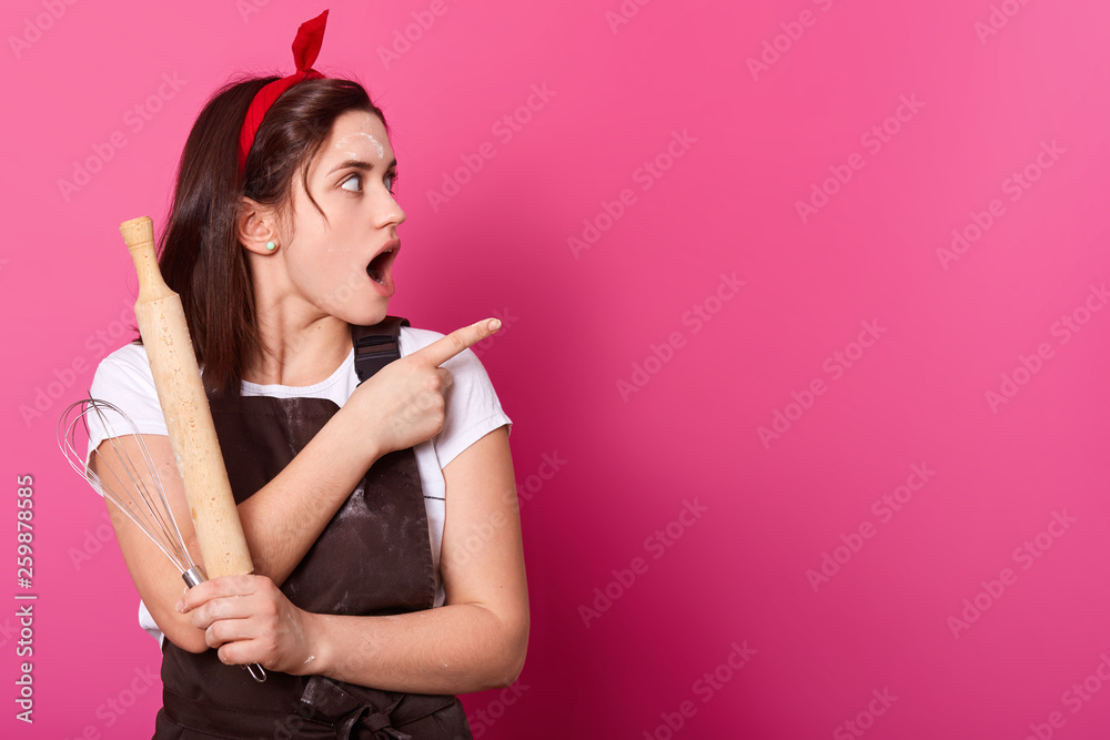 Impressed young girl stands with rolling pin and whisk in her hands, shows direction with forefinger, turns her head on side, opens mouth widely with surprise. Cooking skills development concept. Impressed young girl stands with rolling pin and whisk in her hands, shows direction with forefinger, turns her head on side, opens mouth widely with surprise. Cooking skills development concept.