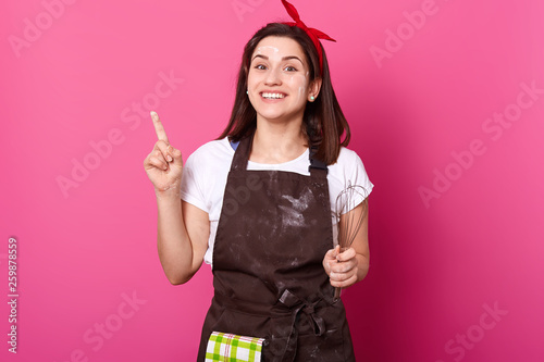 Portrait of adorable young girl holding her finger up. Funny creative female produces new ideas of cooking with enthusiasm. Proffesional baker wears brown apron, white tshirt, bright red headband. photo