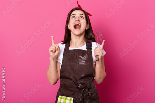 Portrait of brunette energetic girl in dark brown apron smeared with flour, white tshirt and red hair band, stands with her forefingers and head up, holds whisk, looks crazy. Cooking concept. photo