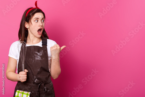 Surprised dark haired woman makes gesture with her thumb. Gifted active baker holds kitchen equipment in right hand, opens her mouth and big eyes widely, wears work clothing. Cooking concept. photo