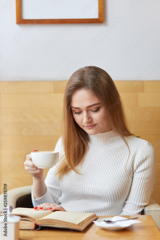 Young model poses sitting in cafe, reading interesting book, holding cup of coffee in right hand, focusing on details of plot. Good looking slender girl feels comfortable and spends her free time. Young model poses sitting in cafe, reading interesting book, holding cup of coffee in right hand, focusing on details of plot. Good looking slender girl feels comfortable and spends her free time.