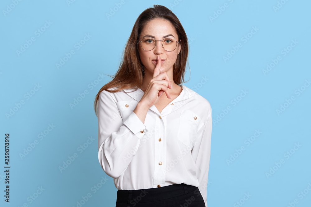 European slender woman shows with gesture to keep in silence, holding forefinger near lips, stands firmly and confidently with her long dark hair down and widely open blue eyes. Copy space for ad. European slender woman shows with gesture to keep in silence, holding forefinger near lips, stands firmly and confidently with her long dark hair down and widely open blue eyes. Copy space for ad.