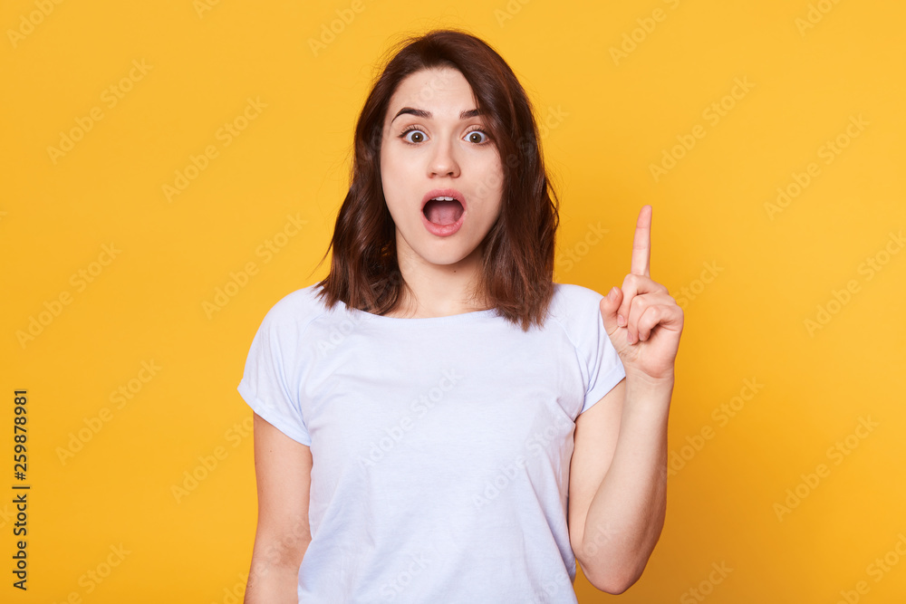 Good looking brunette female dressed casual t shirt, looks in amazement as indicates at something upwards, isolated over yellow background. Charming young woman sees amazing thing up. Copy space. Good looking brunette female dressed casual t shirt, looks in amazement as indicates at something upwards, isolated over yellow background. Charming young woman sees amazing thing up. Copy space.