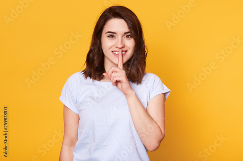 Body language concept. Beautiful smiling young female with happy expression, wears casual t shirt, demonstrates silence gesture, keeps fore finger on lips, isolated on yellow studio background.