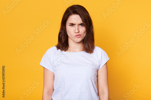 Portrait of angry young woman, frowning over yellow background, looks offened, has unpleasant facial expression, has bad news, brunette female wears casual t shirt poses in photo studio, copy space.