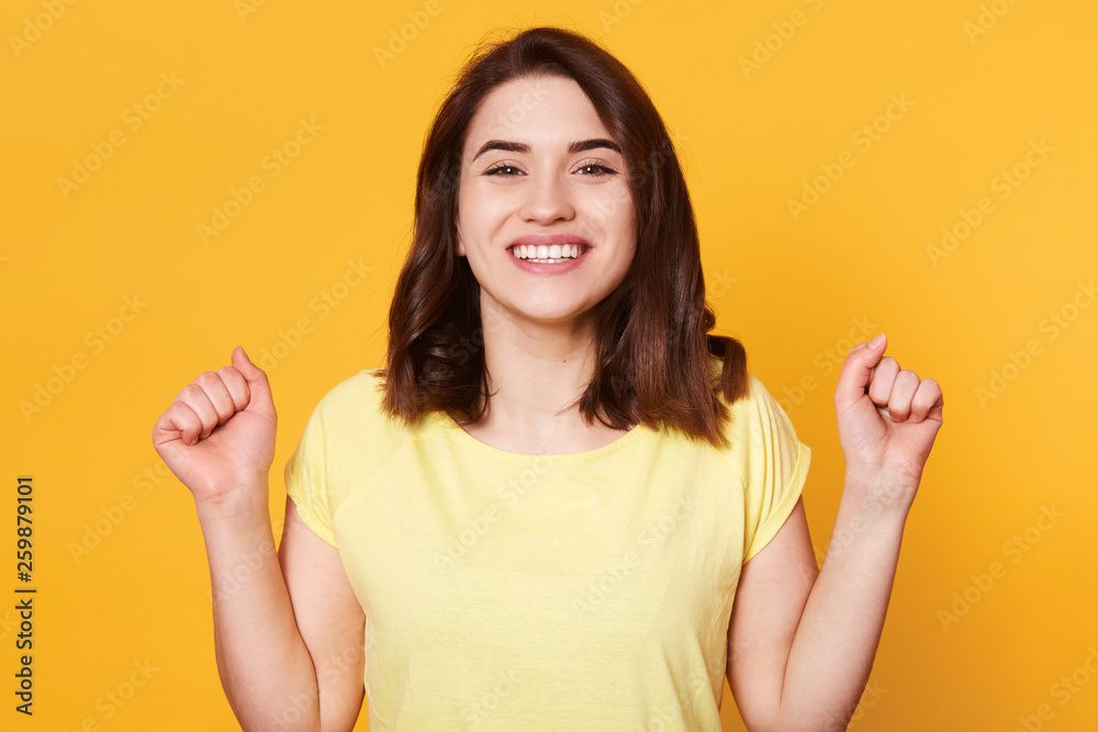 Happy woman dressed casual t shirt, clenching her fists with excitement, celebrating, achieving goal, her dreams come true, posing isolated on yellow background. Success, victory, achievement concept. Happy woman dressed casual t shirt, clenching her fists with excitement, celebrating, achieving goal, her dreams come true, posing isolated on yellow background. Success, victory, achievement concept.