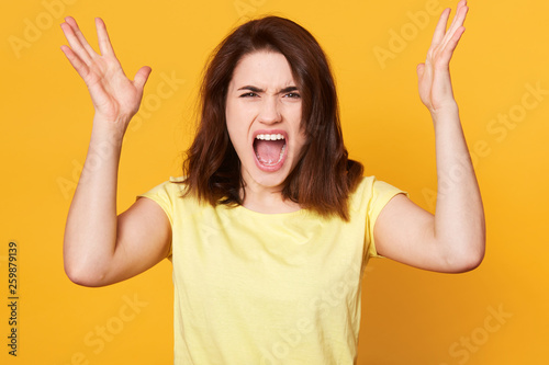 Crying emotional young female clasps hands and looks angrily at camera, has nevous look, has conflict with husband, gestures indoor against yellow background, Human emotions, facial expression concept photo