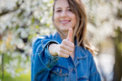 girl in a denim jacket stands near a flowering tree and show Ok hand sign