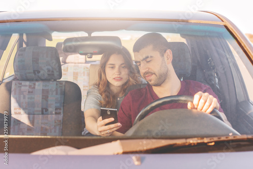 Front view of unshaven male driver sits at wheel and drives his automobile while his wife sits on back seat, holds smart phone and shows her husband online maps, couple find way. Enjoy their weekend. © sementsova321