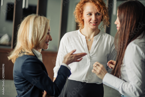 Three positive business women chatting in office lobby. Attractive ladies in white formal shirts, standing and discussing recent seminar. Business coworkers and communication concept.